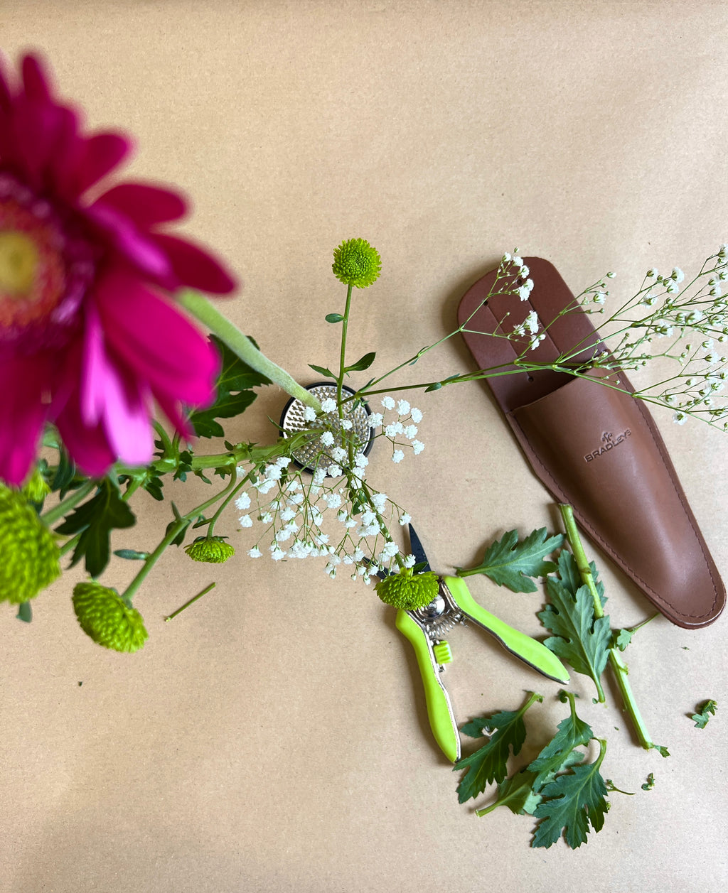 Floral arrangement with a brown leather tool case and green scissors on a beige background